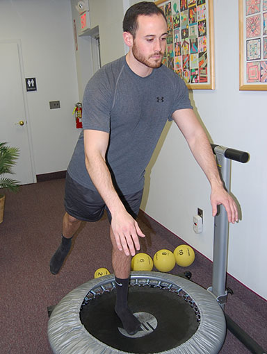 RehabVertical Man in a gray athletic shirt balancing on a mini trampoline in a hallway, with exercise balls and a handrail visible in the background.