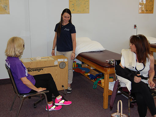 Therapy Therapist supervising a rehab session as a client sits in a chair using a wooden leg-exercise device; another client sits by a treatment table in background.
